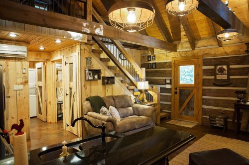 Cozy cabin interior featuring wooden beams, a sofa, and a staircase, with warm lighting and rustic decor.