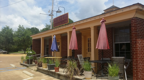 A charming brick building with outdoor seating, colorful umbrellas, and potted plants under a blue sky.
