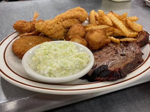 A plate of food featuring fried shrimp, catfish, coleslaw, hushpuppies, crinkle fries, and a slice of brisket.