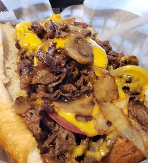 Close-up of a loaded cheesesteak sandwich with sautéed onions, mushrooms, and melted cheese on a hoagie roll.