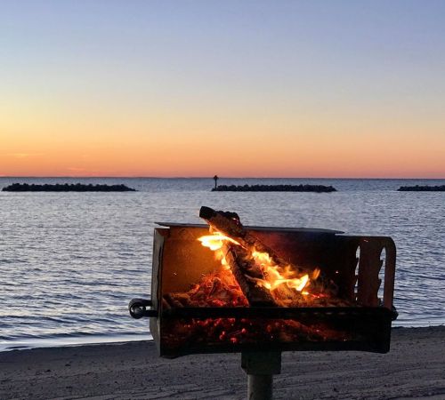 A beachside grill with a fire burning, set against a serene sunset over the water.