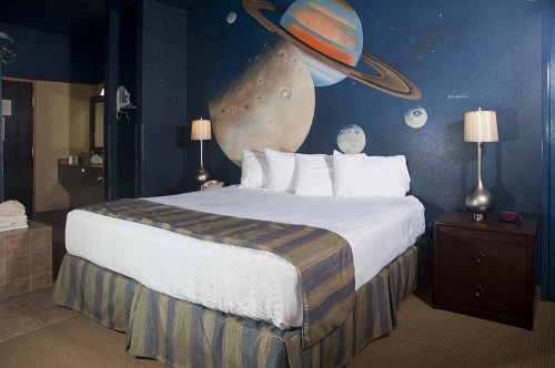 A cozy bedroom with a large bed, two lamps, and a mural of planets and Saturn on the wall.