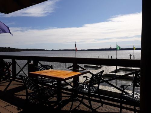 A scenic view of a calm lake from a wooden deck with tables and chairs, featuring colorful flags in the distance.