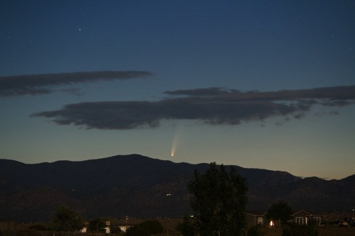 A comet streaks across the night sky above dark mountains, partially obscured by clouds. Stars twinkle in the background.