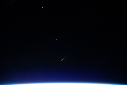 A comet streaks across the dark sky above the Earth's horizon, surrounded by stars.