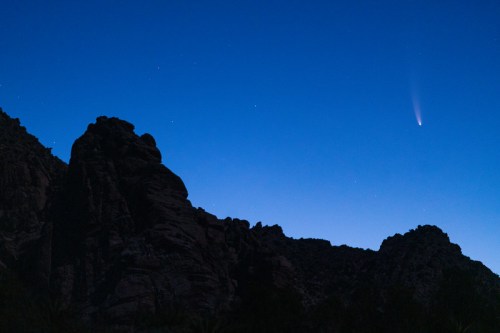 A comet streaks across a twilight sky, silhouetted by rugged mountain peaks and scattered stars.