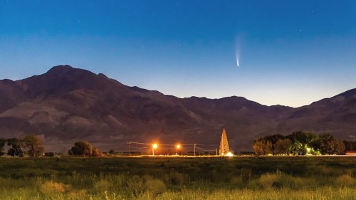 A comet streaks across a twilight sky above mountains, with lights from a nearby field and trees in the foreground.