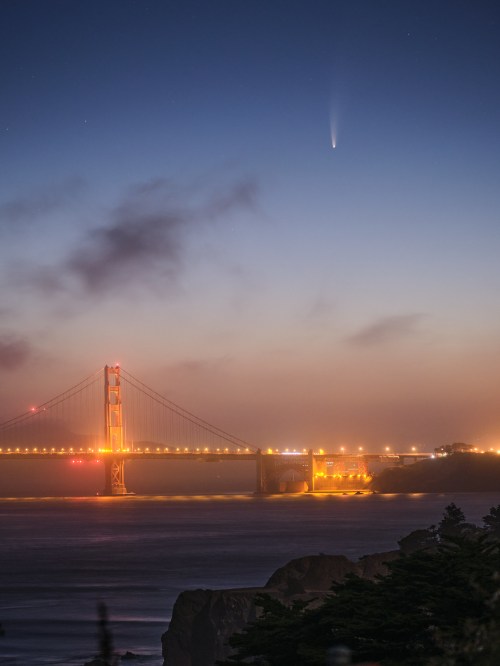 A comet streaks across the twilight sky above the Golden Gate Bridge, illuminated by city lights and ocean waves below.