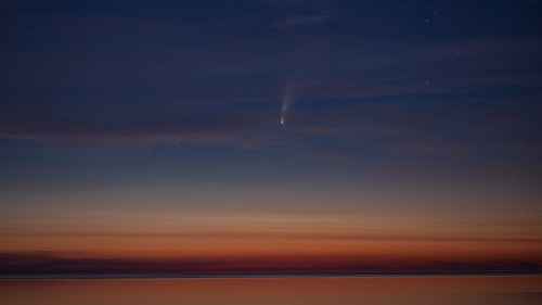 A comet streaks across a twilight sky above a calm ocean, with stars twinkling in the background.