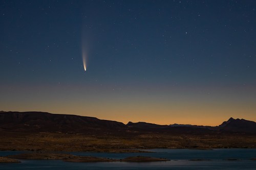 A comet streaks across a starry night sky over a tranquil landscape and calm water.