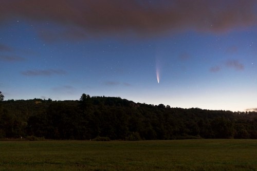 A comet streaks across a twilight sky above a dark, wooded hillside, with scattered stars visible.