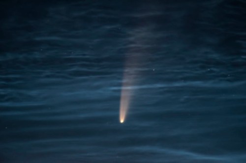 A bright comet streaks across a dark, starry sky, reflecting on calm water below.