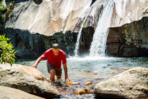 A man in a red shirt wades through a clear stream near a waterfall, surrounded by rocky terrain and greenery.