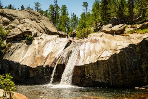 A person stands on a rocky ledge above a waterfall, surrounded by trees and a clear blue sky.