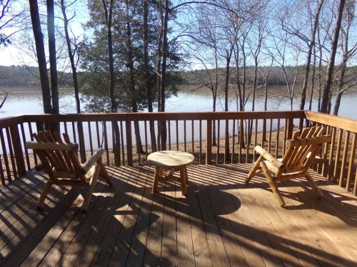 A wooden deck with two chairs and a small table, overlooking a calm lake surrounded by bare trees.