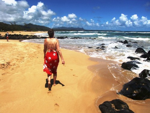 A person walks along a sandy beach, with waves crashing and rocky formations under a bright blue sky.