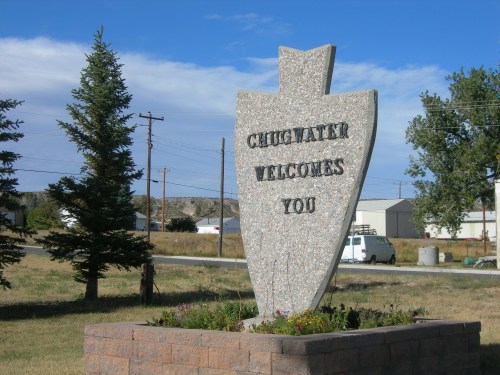 A large stone sign reading "Chugwater Welcomes You" surrounded by grass and trees under a blue sky.