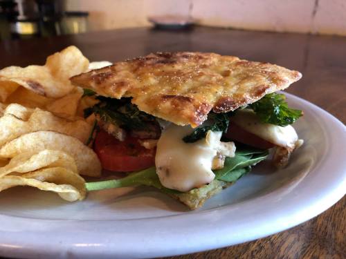 A sandwich with layers of greens, tomatoes, and cheese, served with crispy potato chips on the side.