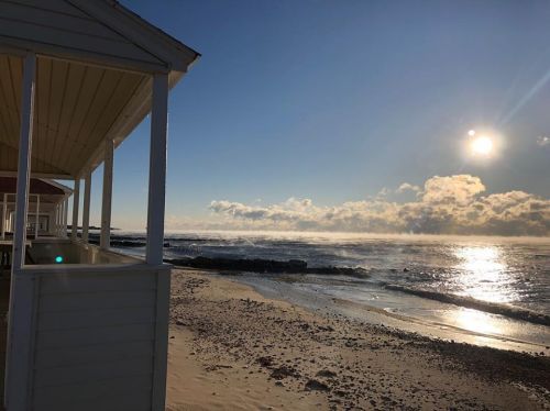 A serene beach scene at sunrise, with gentle waves and a clear sky, framed by white beach cottages.