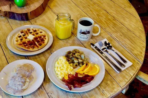A breakfast spread featuring waffles, scrambled eggs, bacon, potatoes, orange slices, and drinks on a wooden table.