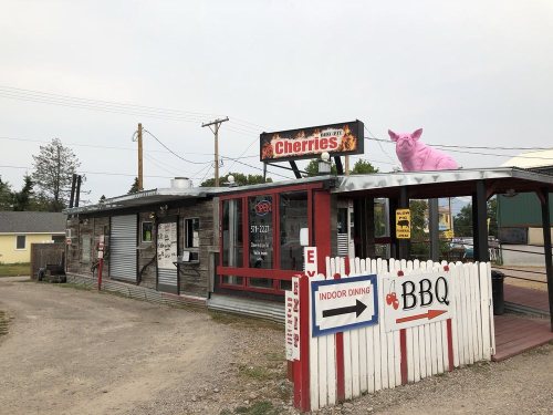 A rustic BBQ restaurant named "Cherries" with a pink pig statue on the roof and outdoor dining signs.