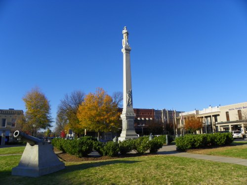 A tall monument with a statue on top stands in a park, next to a cannon and surrounded by trees and buildings.