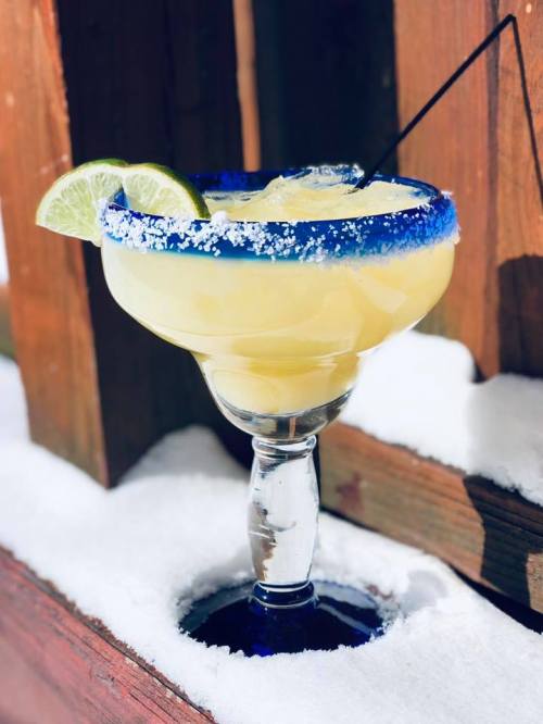 A margarita glass filled with a yellow drink, garnished with lime slices, resting on snow with a blue rim and straw.