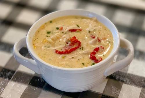 A bowl of creamy chicken soup with shredded chicken, red peppers, and herbs, set on a checkered tablecloth.