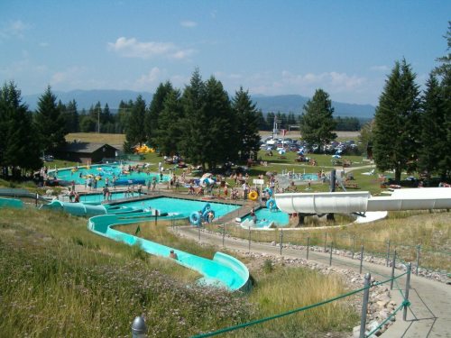 A busy water park with slides, pools, and families enjoying a sunny day surrounded by trees and mountains in the background.