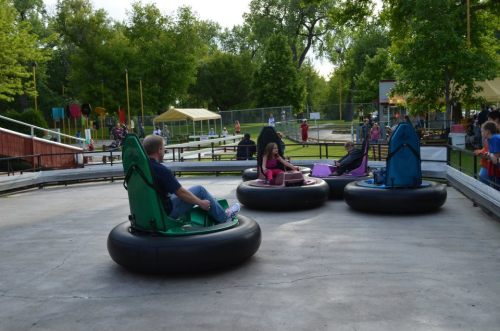 Four people sit in bumper cars on a concrete surface, surrounded by trees and a park setting.