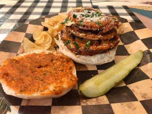 A plate with a stacked sandwich of fried eggplant, topped with sauce and cheese, served with chips and a pickle.