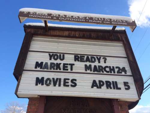 Signboard announcing events: "You Ready? Market March 24, Movies April 5" with snow on top against a blue sky.