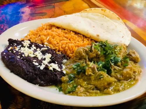 A plate of Mexican food featuring rice, black beans, tortillas, and a green chicken dish garnished with cilantro.