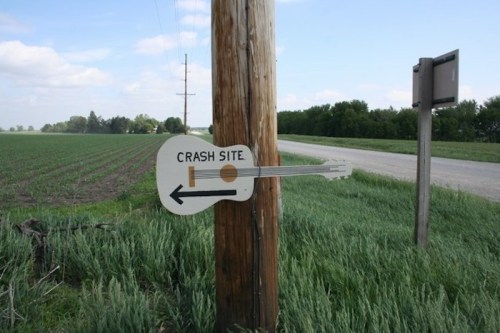 Signpost shaped like a guitar pointing left to a crash site, with a rural landscape and road in the background.