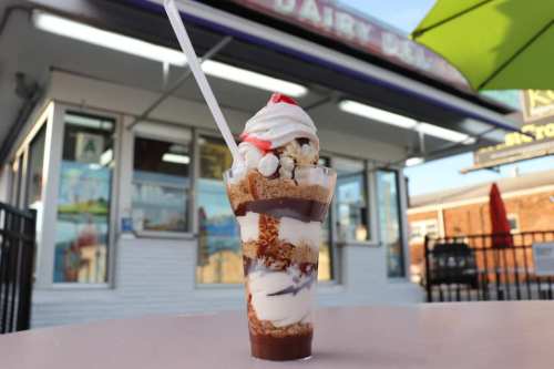 A tall sundae glass filled with layers of ice cream, chocolate sauce, whipped cream, and toppings, set outside a dairy shop.