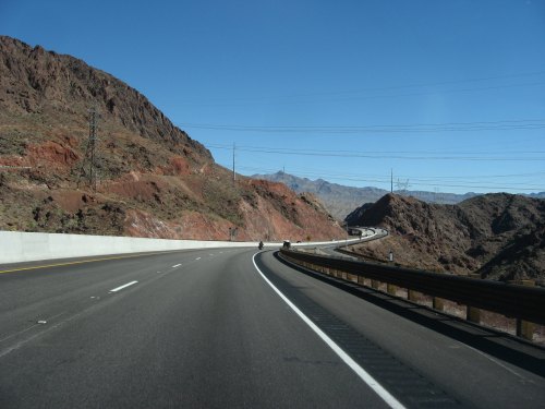A winding highway curves through rocky terrain under a clear blue sky, with mountains in the distance.