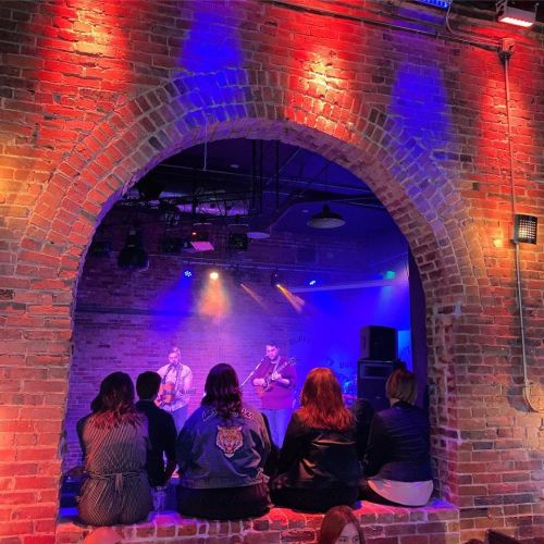 A live music performance in a brick venue, with an audience seated in an archway, illuminated by colorful lights.