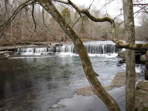 A serene river scene featuring a waterfall, surrounded by bare trees and rocky banks.