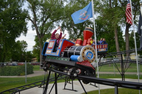 A small roller coaster shaped like a train, with excited riders raising their hands, surrounded by trees and flags.