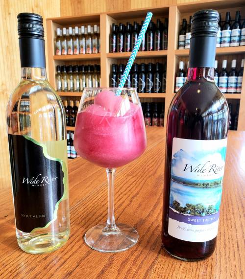 A glass of slushy pink drink with a straw, flanked by bottles of white and red wine on a wooden table.