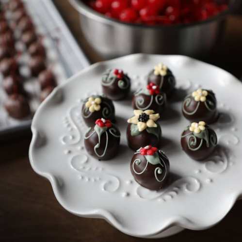 A decorative plate of chocolate truffles topped with flowers and cherries, with a bowl of cherries in the background.