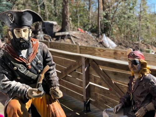 Two pirate statues sit on a wooden deck, one holding a brush and the other wearing an eye patch, surrounded by trees.