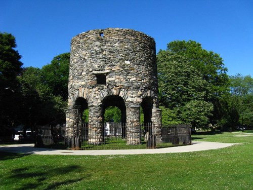 Stone tower with arched openings, surrounded by a black fence, set in a green park under a clear blue sky.