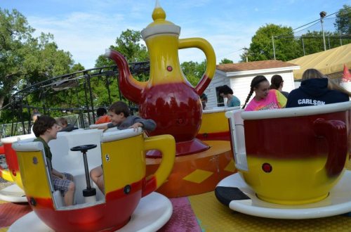Children enjoy a colorful teacup ride at an amusement park, with a large teapot centerpiece in the background.