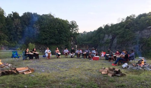 A group of people sitting in a circle around a campfire in a scenic outdoor setting, surrounded by trees and rocks.