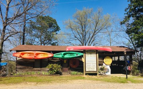 A rustic rental office with colorful kayaks and a canoe displayed outside, surrounded by trees and a clear blue sky.