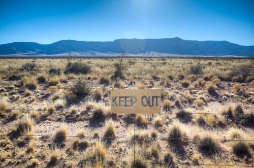 A "Keep Out" sign stands in a dry, grassy landscape with mountains in the background under a clear blue sky.