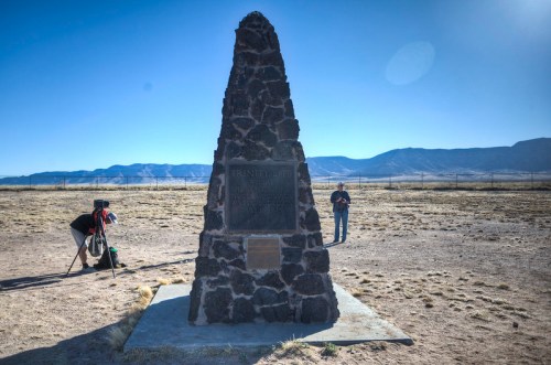 A stone monument stands in a barren landscape, with mountains in the background and two people taking photos nearby.