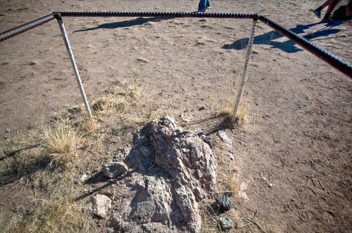 A rocky formation enclosed by metal fencing in a dry, grassy area with people in the background.