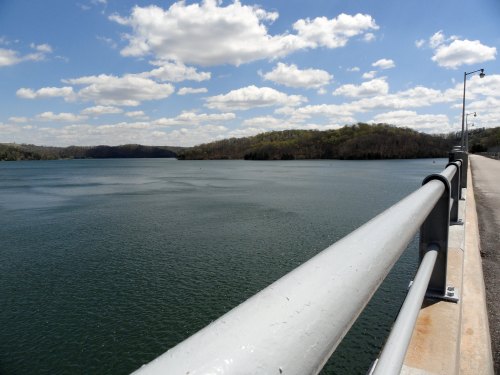A serene lake view with a railing in the foreground, surrounded by green hills and a blue sky with fluffy clouds.